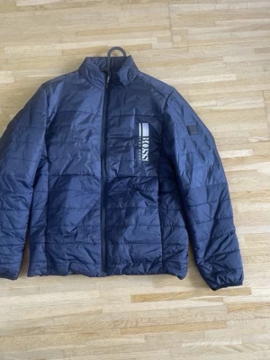 Mörkblå pufferjacka från BOSS - Snygg mörkblå pufferjacka från BOSS med diskret logga på bröstet och ståkrage. Jackan har quiltad design, dragkedja framtill och en modern passform. Perfekt för kalla dagar och enkel att matcha med olika outfits.