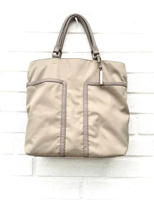 Beige Tommy Hilfiger shoppingväska med läderhandtag - Stilren shoppingväska i beige nylon med matchande läderhandtag och avtagbar axelrem. Väskan har två stora ytterfickor framtill med dekorativa sömmar och en metalltag som detalj. Perfekt för dig som vill ha en enkel men snygg väska till vardagen.