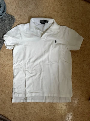 Vit pikétröja från Polo Ralph Lauren - Klassisk vit pikétröja från Polo Ralph Lauren med korta ärmar och den ikoniska loggan broderad på bröstet. Tröjan har krage och knappar vid halsen, tillverkad i mjuk bomull för en bekväm känsla. Säljs för 150kr. Har en liten defekt som skickas i dm på förfrågan. Kontakta mig om du har frågor eller vill lägga bud.