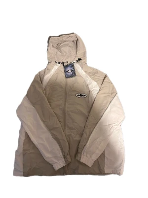Beige vindjacka från Corteiz - Snygg grå vindjacka från Corteiz med huva och dragkedja framtill. Jackan har en sportig look med logga på bröstet den är  perfekt för blåsiga sommar dagar.