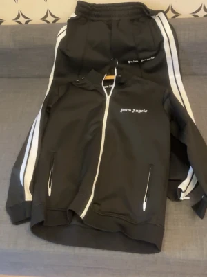 Träningskläder från Palm Angels - Säljer en svart ziptröja från Palm Angels med vita detaljer och dragkedja framtill. Tröjan har vita ränder längs ärmarna och två fickor med dragkedja. Märkets logga är tryckt på bröstet. Perfekt för en sportig och trendig look.