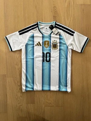 Argentina matchtröja Adidas nr 10 - Säljer en officiell Argentina matchtröja från Adidas med klassiska blå och vita ränder, svarta detaljer och nummer 10 på bröstet. Tröjan har FIFA 2022-märke och AFA-emblem i guld. Tillverkad i lätt och ventilerande material, perfekt för fotboll.