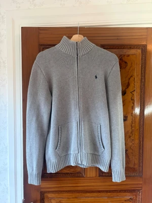Grå ziptröja från Polo Ralph Lauren - Snygg grå stickad ziptröja från Polo Ralph Lauren med hög krage och två fickor fram. Tröjan har ribbade muddar och en liten mörkblå logga broderad på bröstet. Perfekt till lager på lager och enkel att matcha med det mesta.