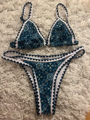 Blommig blå bikini med vita detaljer - Snygg bikini med blå botten och blommigt mönster i vitt och gult. Bikinin har vita och blå triangeldetaljer längs kanterna och smala axelband. Perfekt för stranden eller poolen, ger en fräsch och trendig look.