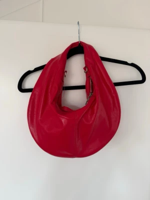 Röd axelväska i skinnimitation - Cool röd handväska i mjuk skinnimitation med rundad, slouchy form. Väskan har dragkedjedetaljer på sidorna och bred axelrem som gör den enkel att bära. Perfekt för dig som vill sticka ut med en färgstark accessoar.