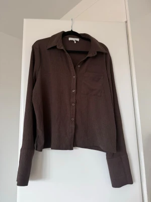 Brun skjorta med lång ärm och ficka från Nakd  - Brun skjorta i ett mjukt tyg med klassisk krage, knappstängning framtill och en bröstficka. Skjortan har långa ärmar och en avslappnad passform, perfekt för en chill och stilren look.
