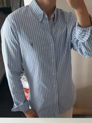 Två skjortor från Ralph Lauren - Säljer två skjortor från Ralph Lauren. En är ljusgrön med klassisk button-down krage och broderad logga, den andra är ljusblå med vita ränder och samma ikoniska logga. Båda har lång ärm, knappar framtill och är i mjuk bomull.