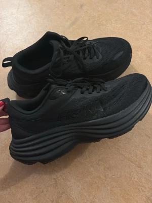 Svarta Hoka Bondi sneakers strl 43 - Säljer ett par helsvarta Hoka Bondi sneakers med chunky sula och mesh-överdel. Skorna har klassisk snörning och diskret logga på sidan. Perfekta för dig som gillar sportig stil och vill ha riktigt bekväma sneakers med extra dämpning. Små size men det passar perfekt till 43 eller 43 1/2 endas provad 