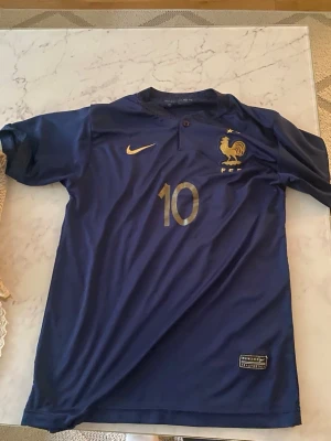 Frankrike fotbollströja Mbappé #10 - Mörkblå officiell Frankrike fotbollströja från Nike med guldiga detaljer, nummer 10 och Mbappé på ryggen. Tröjan har korta ärmar, knapp vid halsen och FFF-logga på bröstet. Perfekt för dig som älskar fotboll och vill stötta landslaget.