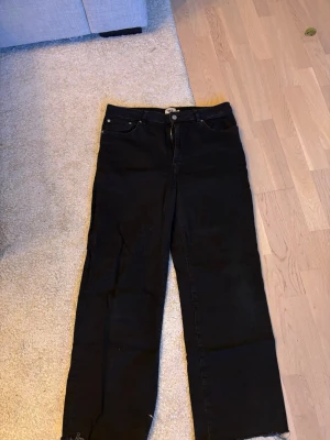 Svarta raka jeans med rå kant - Säljer ett par svarta jeans med rak passform och rå kant nedtill. Jeansen har klassisk femficksdesign, hög midja och stängs med knapp och dragkedja. Perfekta för en avslappnad och trendig look.