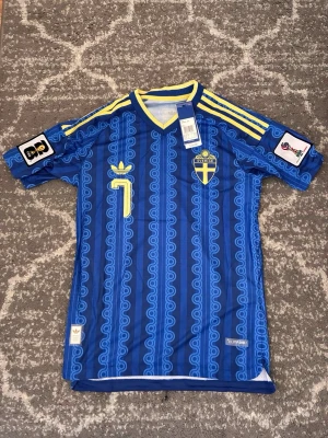 Blå Sverige  fotbollströja Adidas - Retroinspirerad blå fotbollströja från Adidas med Sveriges landslagsemblem och nummer 7 i gult. Tröjan har gula detaljer, tre ränder på axlarna och unikt mönster över hela tyget. Korta ärmar och V-ringad krage. Perfekt för dig som älskar  fotbollsstil.