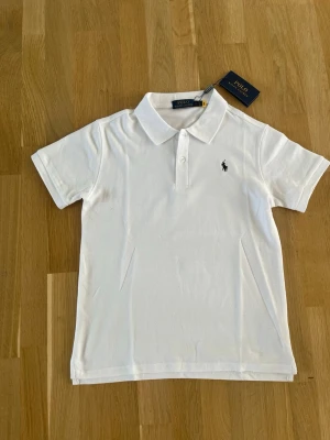 Vit pikétröja från Polo Ralph Lauren - Klassisk vit pikétröja från Polo Ralph Lauren med korta ärmar och krage. Tröjan har knappslå framtill och den ikoniska logotypen broderad på bröstet. Perfekt till jeans eller shorts för en clean och stilren look.