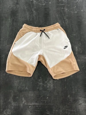 Nike beige och vita shorts med snörning - Nike shorts i beige och vitt med svart snörning i midjan och logga på vänster sida. Tillverkade i mjuk bomull, har elastisk midja och praktisk dragkedjeficka bak. Perfekta för en avslappnad och sportig stil.