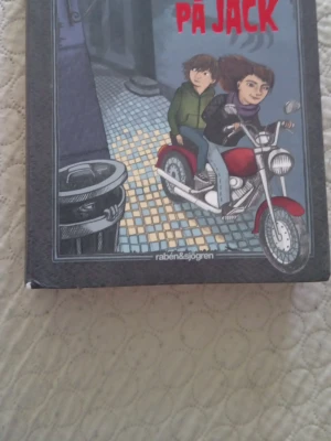 Bok: jakten på Jack - En inbunden bok med titeln 'jaktenpå Jack' från Rabén & Sjögren. Omslaget visar två ungdomar på en röd motorcykel i en stadsmiljö. Boken har hårda pärmar och illustrationer i färg på framsidan.