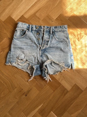 Ljusa högmidjade jeansshorts - Säljer ett par ljusblå, högmidjade jeansshorts med råa, slitna kanter och fransar. Shortsen har klassisk femficksdesign, knappgylf och bälteshällor. Perfekta för varma dagar och ger en avslappnad vibe.