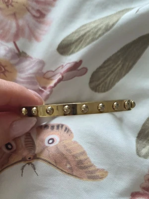 Guldigt armband BY JOLIMA - Snyggt armband i guldton från BY JOLIMA, tillverkat i rostfritt stål. Armbandet har en stilren, rundad form och är dekorerat med små runda detaljer längs hela bandet för en cool och modern look.