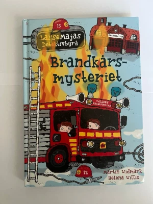 Brandkårsmysteriet - LasseMajas Detektivbyrå - WBrandkårsmysteriet är en illustrerad barnbok från serien LasseMajas Detektivbyrå, skriven av Martin Widmark och illustrerad av Helena Willis. Omslaget är färgglatt med en röd brandbil, eldslågor och blå bakgrund. Perfekt för unga deckarfans som gillar spännande mysterier.