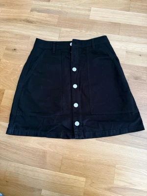 Svart kjol från Tommy Jeans - Svart kjol från Tommy Jeans med vita knappar längs hela framsidan. Stl. XS