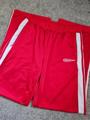 Röda Sportiga Trackpants – Ninetytwo Club (New Yorker) - Säljer ett par snygga och klassiska trackpants i en klar röd färg. Byxorna har vita revärer längs sidorna med röda detaljer och märket "Ninetytwo" broderat på framsidan.