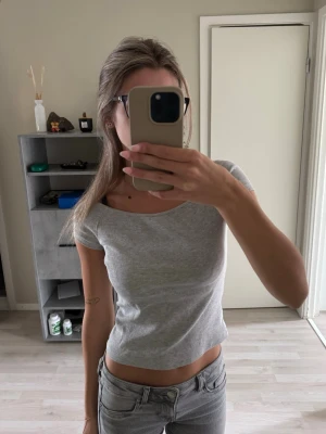 Ljusgrå ribbad croppad topp - Säljer en ljusgrå ribbad croppad topp med korta ärmar och rund halsringning. Toppen har en slim passform och är tillverkad i ett mjukt bomullsmaterial som sitter snyggt på kroppen. Perfekt att styla med jeans eller kjol. Säljer då den inte kom till användning.