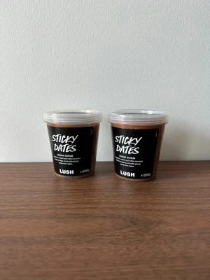 Lush Sticky Dates Sugar Scrub - Sticky Dates Sugar Scrub från Lush i två burkar om 290g vardera. Skrubben har en brun färg och kommer i genomskinliga plastburkar med svart etikett och vitt tryck. Perfekt för exfoliering och att ge huden en mjuk och fräsch känsla. Bägge förpackningarna är oöppnade 