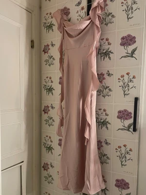 Ljusrosa långklänning med volanger - Elegant ljusrosa långklänning med breda axelband och volangdetaljer längs sidorna. Klänningen har en mjuk, glansig yta och en feminin siluett som faller snyggt. Perfekt för speciella tillfällen där du vill känna dig extra fin. Helt ny ifrån cider som endast är provad.