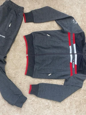 Mörkgrått loungewear-set Tommy Hilfiger - Mörkgrått loungewear-set från Tommy Hilfiger med röda och vita detaljer. Setet består av en ziptröja med fickor och matchande byxor med resår i midjan. Tröjan har röda och vita ränder över bröstet och logga på både tröja och byxa. Mjukt material, perfekt för chill dagar.