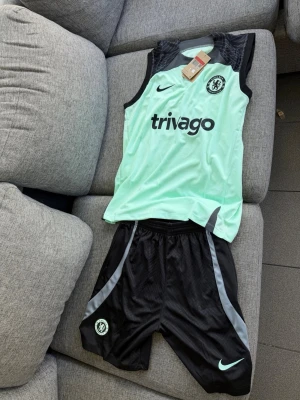 Chelsea träningsset från Nike - Chelsea FC träningsset med ärmlös mintgrön tröja och svarta shorts. Tröjan har svart axelparti, tryckt klubbmärke och Trivago-logga. Shortsen har klubbmärke och Nike-logga samt ljusgröna detaljer. Perfekt för fotbollsträning eller gym. Material: polyester.