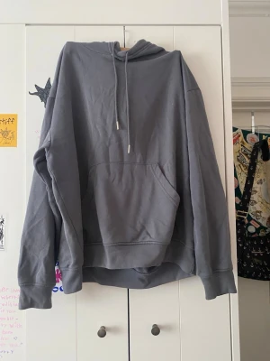 Oversized grå hoodie med ficka - En oversized grå hoodie med stor magficka och dragsko i huvan. Hoodien har långa ärmar och breda muddar vid ärmslut och nederkant. Perfekt för en chill och avslappnad stil. Materialet är mjukt och bekvämt, troligtvis bomullsblandning.
