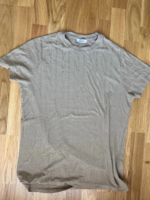 Beige t-shirt från Valentino - Säljer en beige t-shirt från Valentino med diskret mönster i tyget och rund halsringning. Tillverkad i Italien och har en klassisk passform med korta ärmar. Perfekt för dig som gillar stilrena och exklusiva plagg.