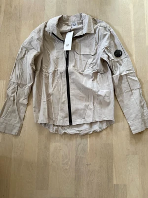 Beige overshirt från C.P. Company - Snygg beige overshirt från C.P. Company med svart dragkedja, klassisk krage och bröstficka. Ikonisk linsdetalj på ärmen och lätt material som ger en modern look. Perfekt lager-på-lager-plagg för dig som gillar streetwear och stilrena detaljer.