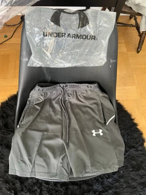 Grå träningsshorts från Under Armour - Snygga grå träningsshorts från Under Armour med elastisk midja och snörning. Shortsen har logga på vänster ben och är tillverkade i ett lätt och snabbtorkande material, perfekta för gymmet eller löprundan.