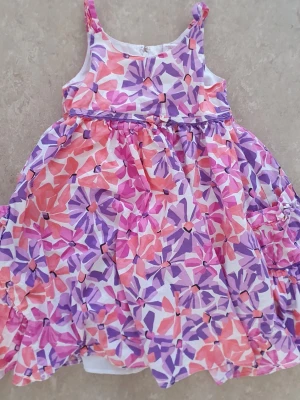 Klänning - Söt klänning med färgglatt blommönster i lila, rosa och orange toner. Klänningen har smala axelband och en utställd kjol, perfekt för varma dagar. Tillverkad i mjuk bomull som känns skön mot huden.