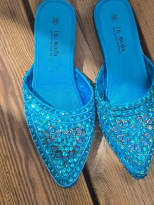 Blå glittriga ballerinaskor La Moda - Säljer ett par blå ballerinaskor från La Moda med spetsig tå och öppen häl. Skorna är täckta av glittriga stenar och pärlor i olika former och har en elegant, skimrande finish. Perfekta för dig som vill sticka ut med färg och bling.