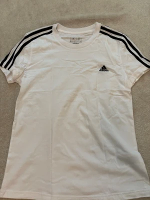 Vit Adidas t-shirt med svarta ränder - Klassisk vit t-shirt från Adidas med svarta ränder längs axlarna och det ikoniska Adidas-loggan på bröstet. T-shirten har rund hals och korta ärmar, perfekt för en sportig och avslappnad stil.