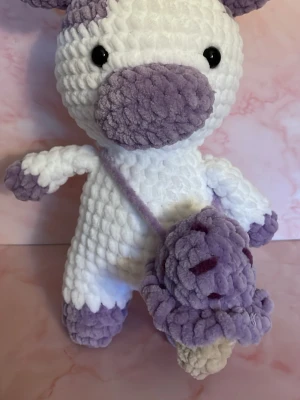 Virkad mjukisdjur med glassväska - Unikt handgjort mjukisdjur i tjockt, fluffigt garn. Djuret är vitt och lila med stora svarta ögon och har en liten axelväska formad som en lila glass med beige strut. Perfekt som dekoration eller present till någon som gillar söta accessoarer.