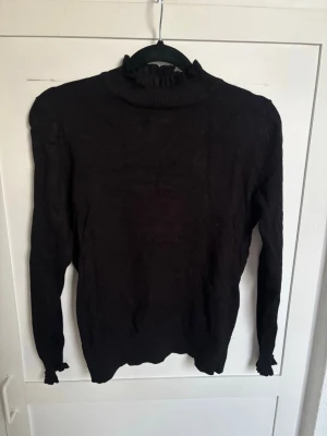 Svart stickad polotröja med volang - Säljer en svart stickad polotröja med volangdetaljer vid krage och ärmslut. Tröjan har lång ärm och är figurnära i modellen. Perfekt för dig som gillar en enkel men ändå trendig look.