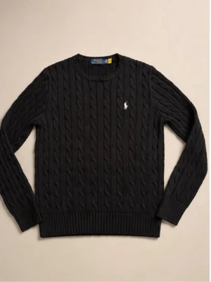Svart kabelstickad tröja Polo Ralph Lauren - Kabelstickad svart tröja från Polo Ralph Lauren med rund halsringning och den klassiska vita broderade loggan på bröstet. Tröjan har ribbade muddar vid ärmslut och nederkant, perfekt för en stilren och tidlös look. Snygg till sommaren och stilren