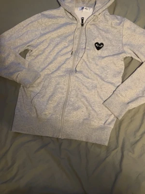 Grå hoodie från Comme des Garçons Play - Snygg grå zip-hoodie från Comme des Garçons Play med det ikoniska svarta hjärtat med ögon på bröstet och en stor version på ryggen. Mjuk bomullskvalitet, dragsko i huvan och ribbade muddar. Perfekt för en chill och trendig look.