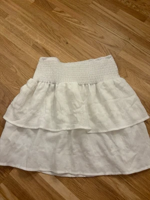 Vit volangkjol - Söt vit kjol med bred smock i midjan och dubbla volanger. Kjolen har en luftig och lätt känsla, perfekt för varma dagar. Passar till både sneakers och sandaler för en avslappnad look.