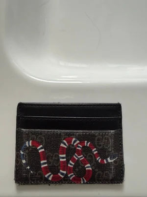 Plånbok - Snygg korthållare från Gucci i svart och grått med klassiskt GG-mönster och en cool röd, vit och svart orm på framsidan. Tillverkad i canvas och läder, har flera fack för kort. Perfekt accessoar för dig som gillar lyxiga detaljer.