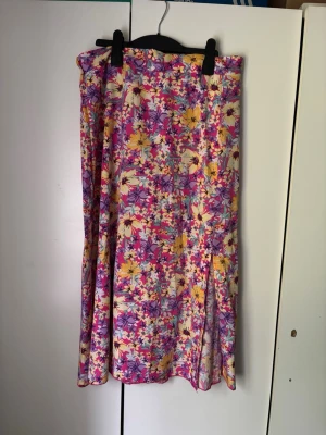 Blommig långkjol i färgglada toner - Färgglad långkjol med blommigt mönster i rosa, lila, gul och vit. Kjolen har en flowy passform och är tillverkad i mjuk polyester med lite stretch för extra komfort. Perfekt för dig som vill sticka ut med en somrig och lekfull stil.