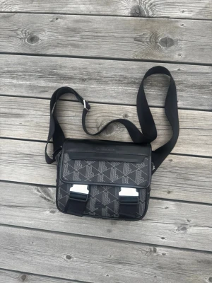 Lacoste bag 🐊 - * Original Lacoste-design med svart krokodillogga. * Snygga grå detaljer som ger väskan en modern och exklusiv känsla. * Justerbar axelrem med spänne för att enkelt anpassa längden efter önskemål. * Rymlig och funktionell för vardagliga tillhörigheter. * Lätt och bekväm att bära. * Perfekt för både vardag och resor. * Mycket väl omhändertagen och utan synliga tecken på slitage.  Skick:  * Nyskick – mycket sparsamt använd.