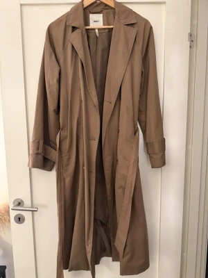 Beige trenchcoat från OBJECT - Stilren beige trenchcoat från OBJECT med klassisk krage, knytskärp i midjan och långa ärmar med slejfdetaljer. Rak passform och två sidofickor. Perfekt för lager på lager och en tidlös look.