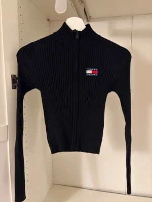 Svart ribbstickad tröja Tommy Jeans - Snygg svart cropped ribbstickad och figurnära cardigan från Tommy Jeans med dragkedja och turtle neck. Tröjan har lång ärm och en broderad logga på bröstet i rött, vitt och blått. 