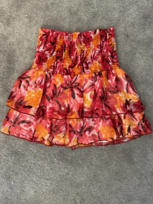 Blommig volangkjol från Gina Tricot - Kort blommig kjol från Gina Tricot i färgerna röd, orange och gul. Kjolen har smock i midjan och dubbla volanger för en luftig och somrig känsla. Perfekt för dig som vill ha en färgstark och trendig look.