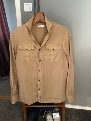 Loro Piana Linen Overshirt - Loro Piana Linne Militärinspirerad Overshirt  Perfekt sommarklädesplagg från Loro Piana. Tillverkad i mjuk och andningsbar italiensk linne i en snygg khaki/beige färg. Klassiska militära detaljer med två fickor med luckor på bröstet, knäppning fram och justerbara ärmar.  Storlek M men passar även L bra tack vare den avslappnade passformen. Kan bäras öppen som lätt jacka eller knäppt.  Skicka ett meddelande vid frågor