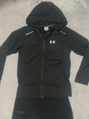 Svart Under Armour tracksuit  - Snyggt svart träningsset från Under Armour med dragkedja på jackan och matchande byxor med dragsko i midjan. Setet har diskreta loggor på både jacka och byxor samt praktiska fickor. Perfekt för gymmet eller löprundan.