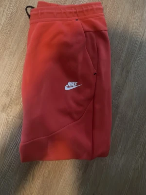Röda Nike tech mjukisbyxor - Säljer ett par röda mjukisbyxor från Nike med vit logga på benet. Byxorna har resår i midjan, muddar vid bensluten och dragkedjeförsedd ficka. Tillverkade i mjukt material som känns skönt mot huden. Perfekta för chill dagar eller träning.