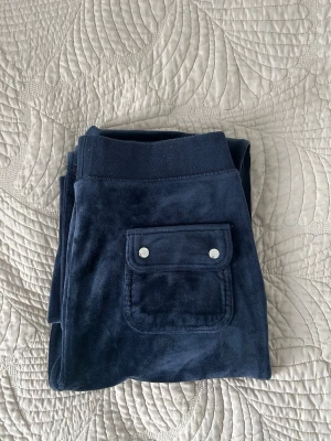 Mörkblå velour mjukisbyxor juicy couture - Mörkblå velour mjukisbyxor juicy couture i mjuk velour med bred resår i midjan och en ficka bak med två silvriga knappar. Perfekta för chill dagar och har en klassisk rak passform. Materialet är lent och ger en skön känsla mot huden.